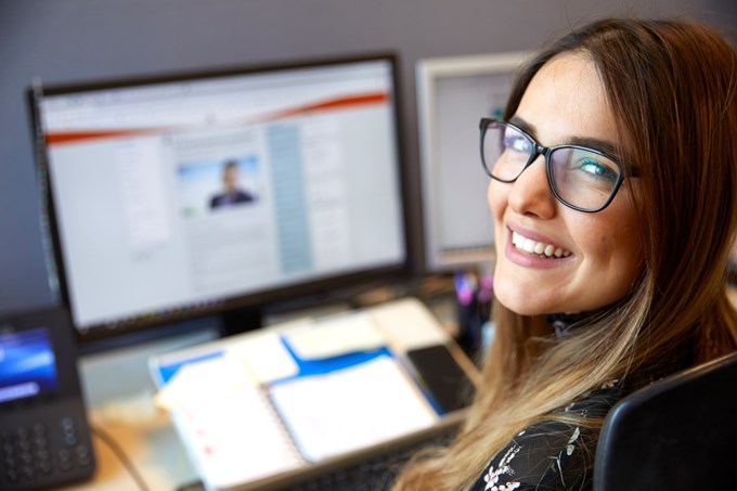 En kvinna med mörkt hår och glasögon sitter framför en dataskärm. Hon vrider huvudet snett bakot och tittar leende in i kameran.