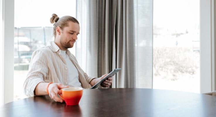 Man som sitter i profil vid ett köksbord. I ena handen håller han en surfplatta och i andra handen en orange kaffekopp.