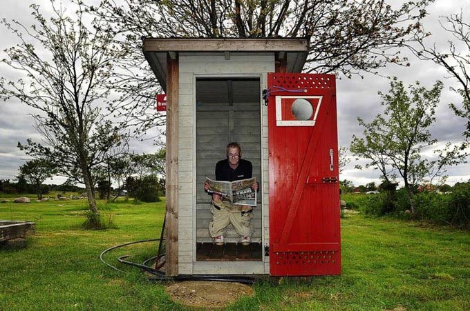 person som sitte ri ett rött utedass och läser tidningen. Dasset står på en grön gräsmatta