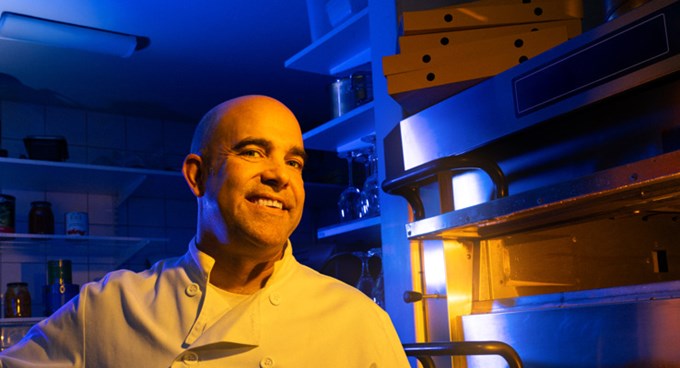 Person i arbetskläder står i ett restaurangkök bredvid en pizzaugn med hyllor och förvaringslådor i bakgrunden.