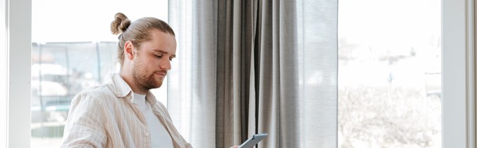 Man med knut i håret sitter vid ett bord och håller i en mobiltelefon framför ett stort fönster.