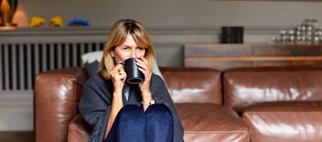 Kvinna inlindad i en filt dricker kaffe i en soffa.